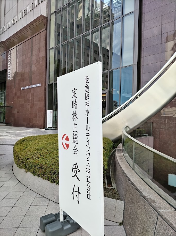 梅田芸術劇場が入る「ホテル阪急インターナショナル」の茶色いモダンな建物の外観を背景に、屋外の敷地内に設置された「阪急阪神ホールディングス株式会社 定時株主総会 受付」と書かれた白い縦型の案内看板と、進行方向を示す赤い矢印マーク。