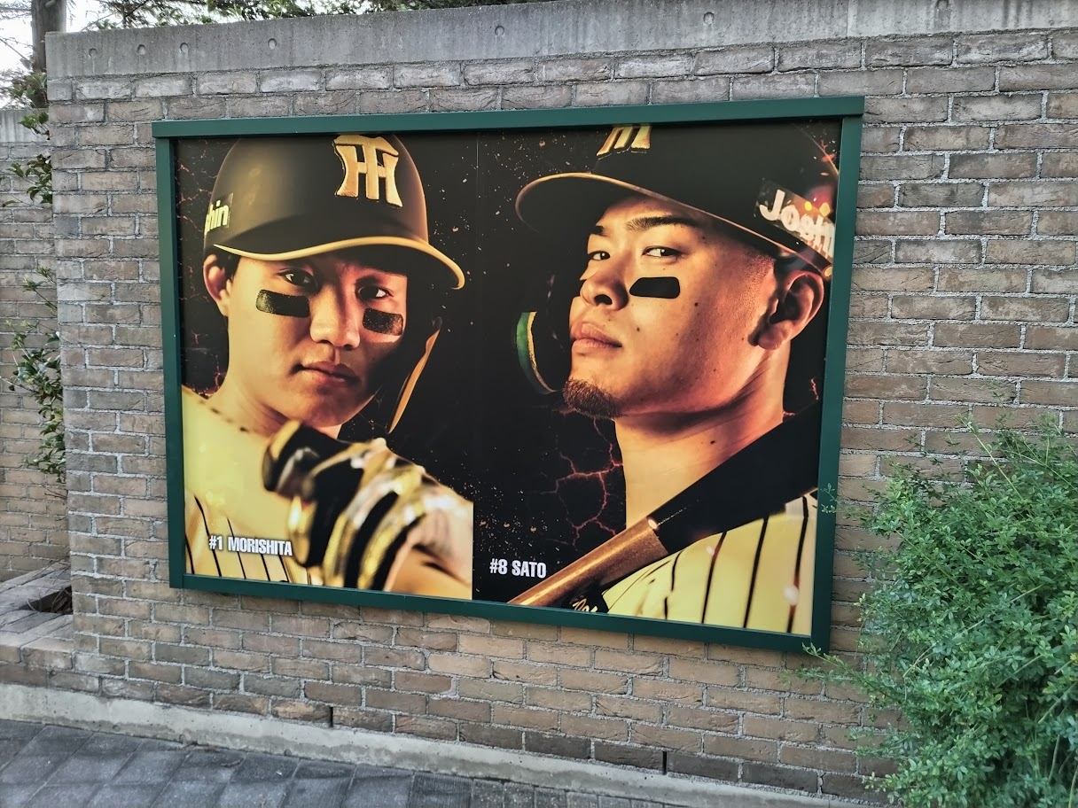阪神甲子園球場のレンガ壁に飾られた、森下翔太選手と佐藤輝明選手の凛々しい大型ビジュアルパネルの写真。二人がアイブラックをつけてバットを構えた力強い表情が描かれている。