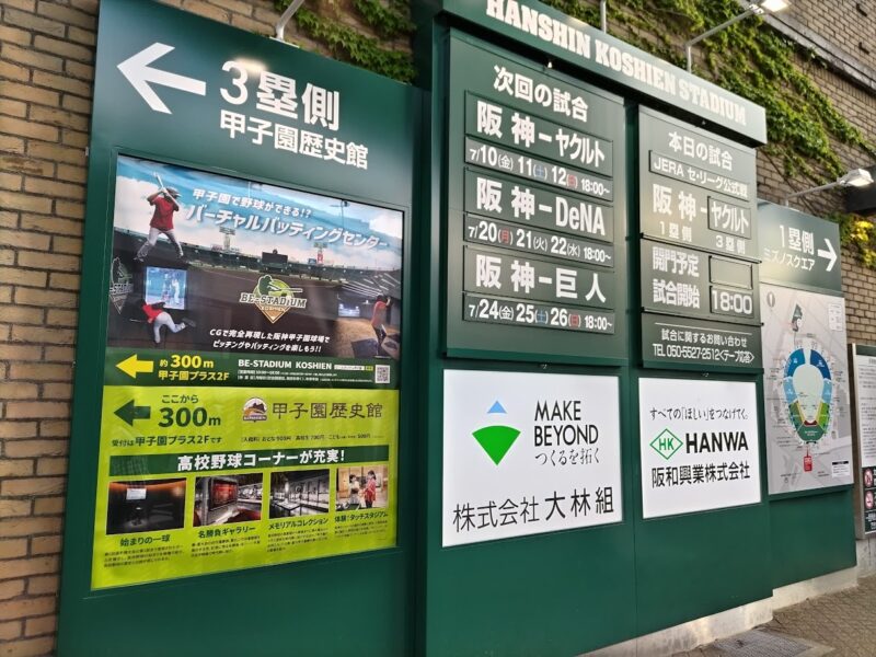 阪神甲子園球場の外壁に設置された試合案内板の写真。本日の試合として「JERA セ・リーグ公式戦 阪神対ヤクルト」のカードと試合開始18:00の予定が掲示されており、左側には甲子園歴史館やバーチャルバッティングセンターの案内ポスターが並んでいる。