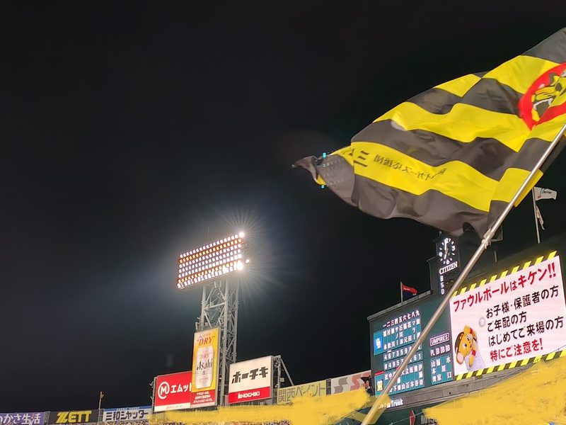 夜の甲子園球場外野スタンドから見上げる、風になびく黄色と黒の応援旗とカクテル光線の照明灯