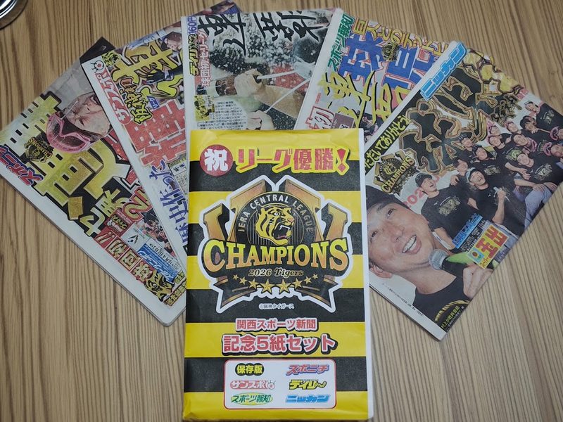 「祝リーグ優勝！」と書かれた黄色と黒のタイガースカラーのパッケージを中心に、木目のテーブルの上に並べられた関西スポーツ新聞の優勝記念5紙セット（サンスポ、スポニチ、スポーツ報知、デイリー、ニッカン）。